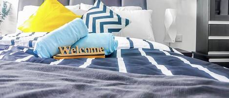 A neatly made bed with a striped bedspread, a 'Welcome' sign, and decorative pillows.