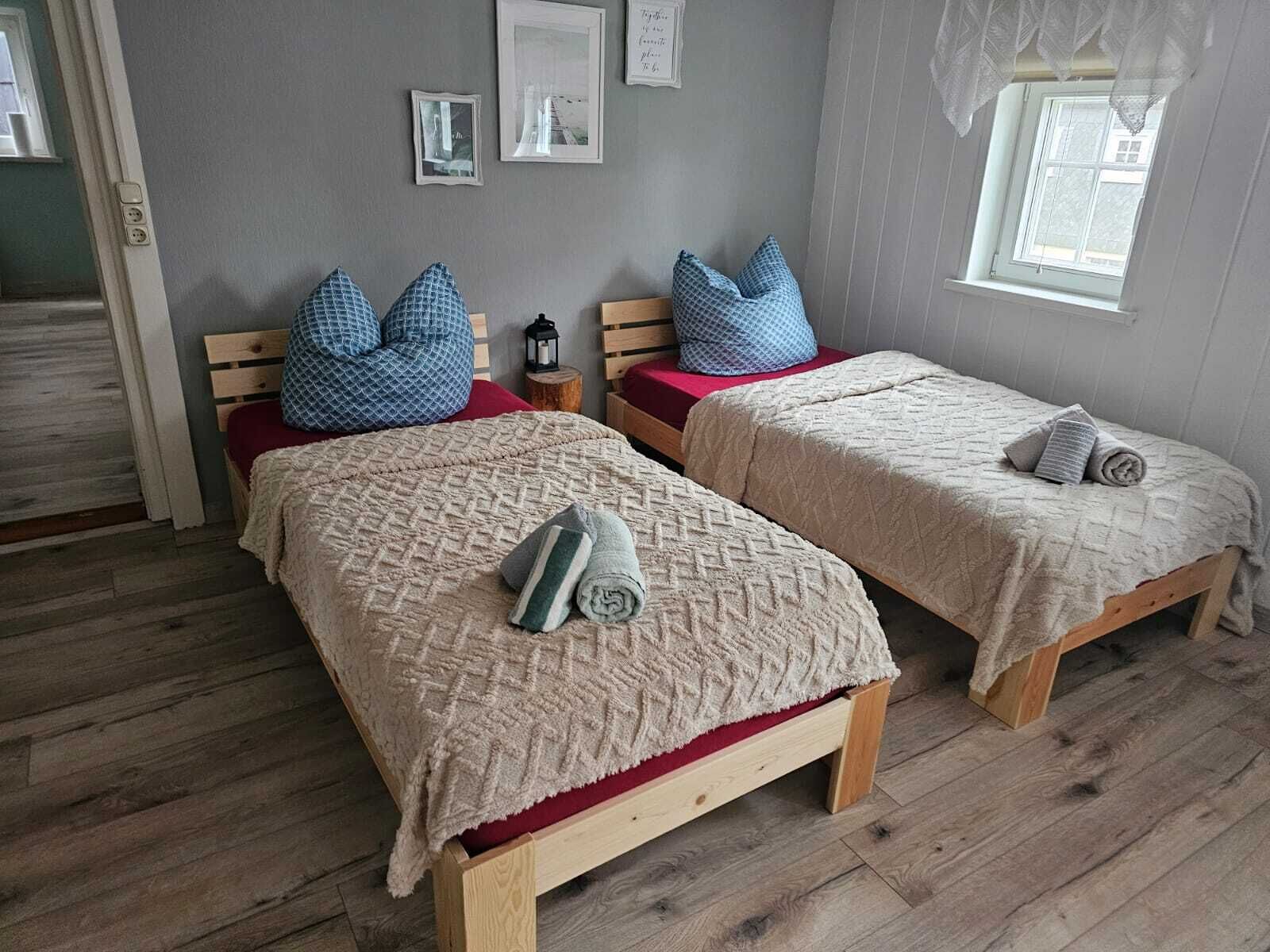 A room with two wooden beds, each with a red mattress and a beige blanket. There are blue pillows on the beds, and a small wooden stool with a lantern beside one bed.