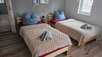 A room with two wooden beds, each with a red mattress and a beige blanket. There are blue pillows on the beds, and a small wooden stool with a lantern beside one bed.