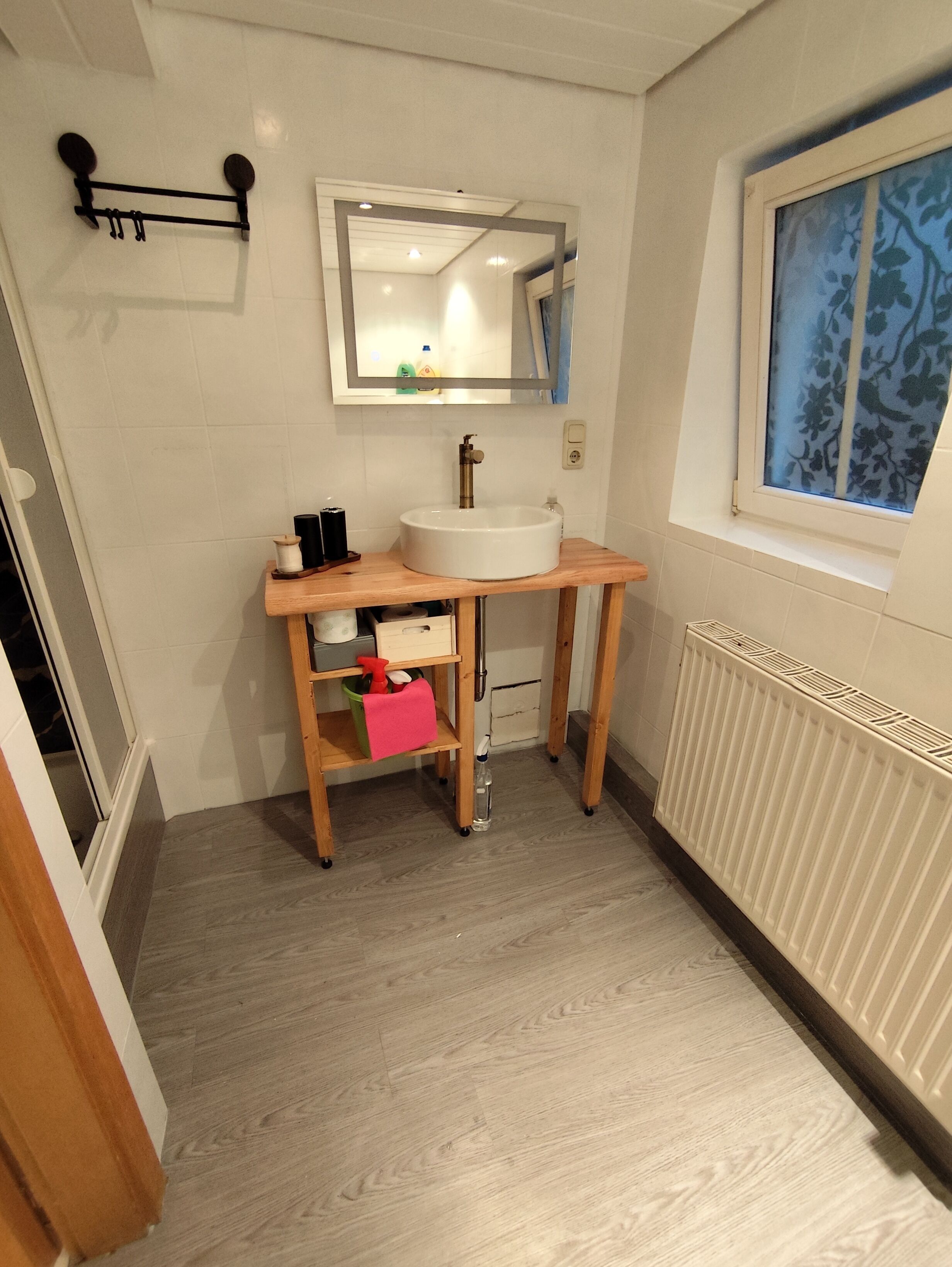 Une salle de bain avec un meuble-lavabo en bois, un lavabo blanc, un miroir et une fenêtre ornée d’un motif floral.