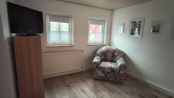 A room with wooden flooring, a radiator, two windows with blinds, a television on a cabinet, and a floral-patterned armchair.