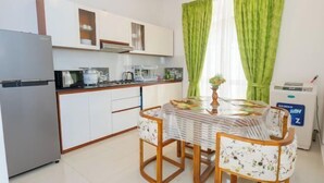 A compact kitchen with white cabinets, a refrigerator, a coffee machine, and a table with chairs. - The Hideout - 2Br Villa Near Kahathuduwa Interchange (Polgasowita)