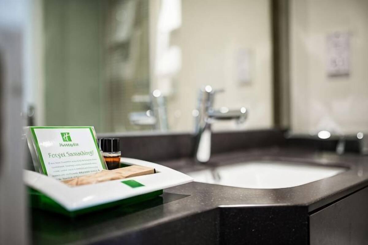 Un baño con lavamanos, espejo y una bandeja con un letrero verde y una botella.