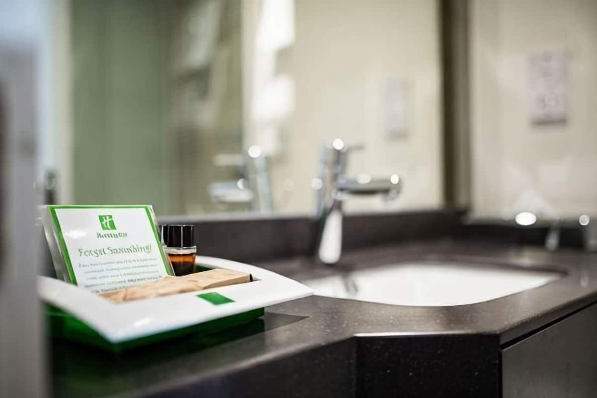 Une salle de bain avec un lavabo, un miroir et un plateau sur lequel se trouvent un panneau vert et une bouteille.