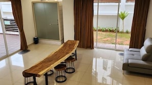 A modern living room with a long wooden table, four stools, a grey sofa, and large windows with curtains. - Zinnia Residence Tanah Rata (Tanah Rata)