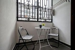 Een klein balkon met twee metalen stoelen, een tafeltje en een raam met decoratieve ijzeren spijlen.