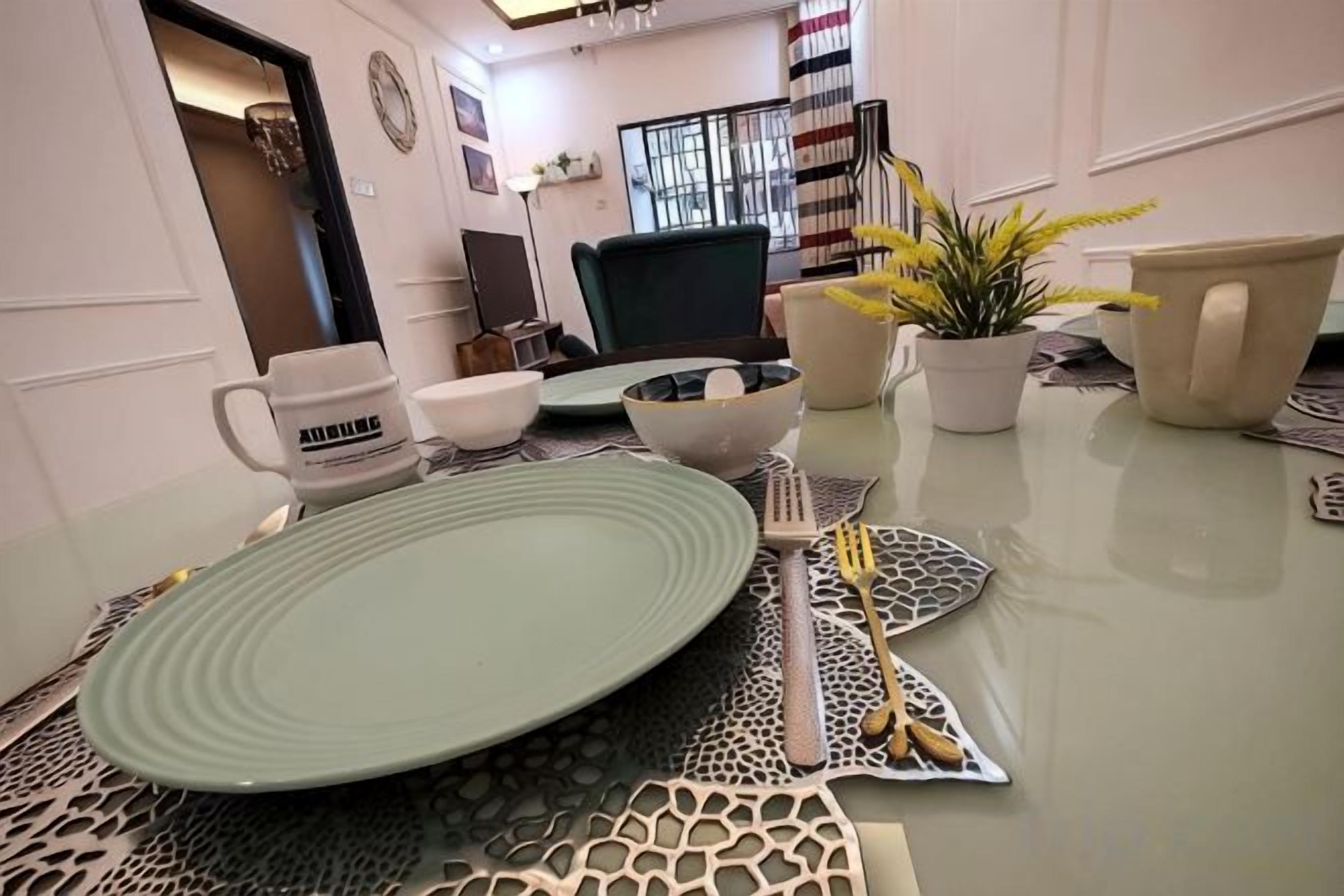 Una mesa de comedor con un plato verde, cuencos blancos y un tenedor. Se ven macetas con plantas y una silla.