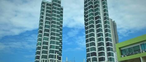 Two tall, modern high-rise buildings with a glass facade, a covered walkway, and a green building with signage.