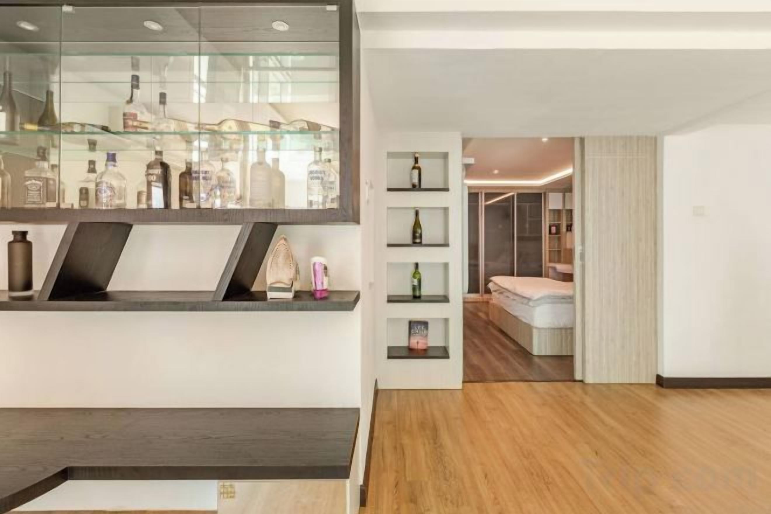 A hotel room with a wooden floor, a glass display cabinet with bottles, and a bed with a white headboard.