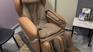 Un fauteuil de massage doté d’un siège et d’accoudoirs rembourrés, placé dans une pièce au sol en marbre et où se trouve une petite table avec un panneau.