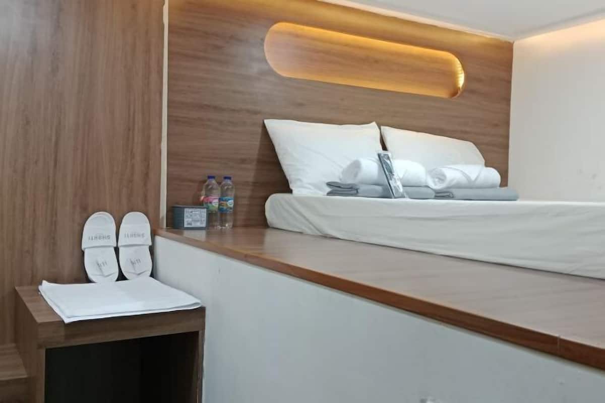 A modern hotel room with a wooden headboard, a neatly made bed, a small bedside table with slippers, and a wall-mounted light fixture.