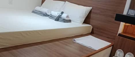Una cama bien hecha con almohadas y toallas dobladas, una mesita de noche de madera con un bloc de notas blanco, y un par de pantuflas en el suelo.