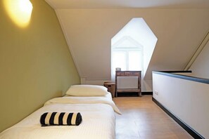 A small, well-lit room with a sloped ceiling, a bed with a striped pillow, a small table, and a window. - Luxury Thai Design Home – Chinatown Self Check In (Bangkok)