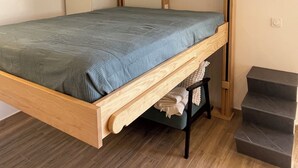 A bunk bed with a wooden frame and a blue mattress. There is a small wooden stool under the bed and a set of stairs to the right.