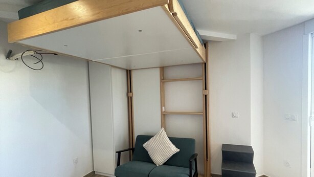 A room with a bunk bed, a sofa with a pillow, a staircase, and a wooden floor.