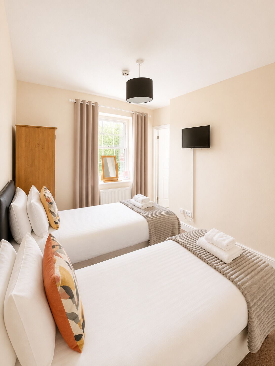 A hotel room with two beds, a wooden wardrobe, a window with curtains, a flat-screen TV, and a ceiling light.