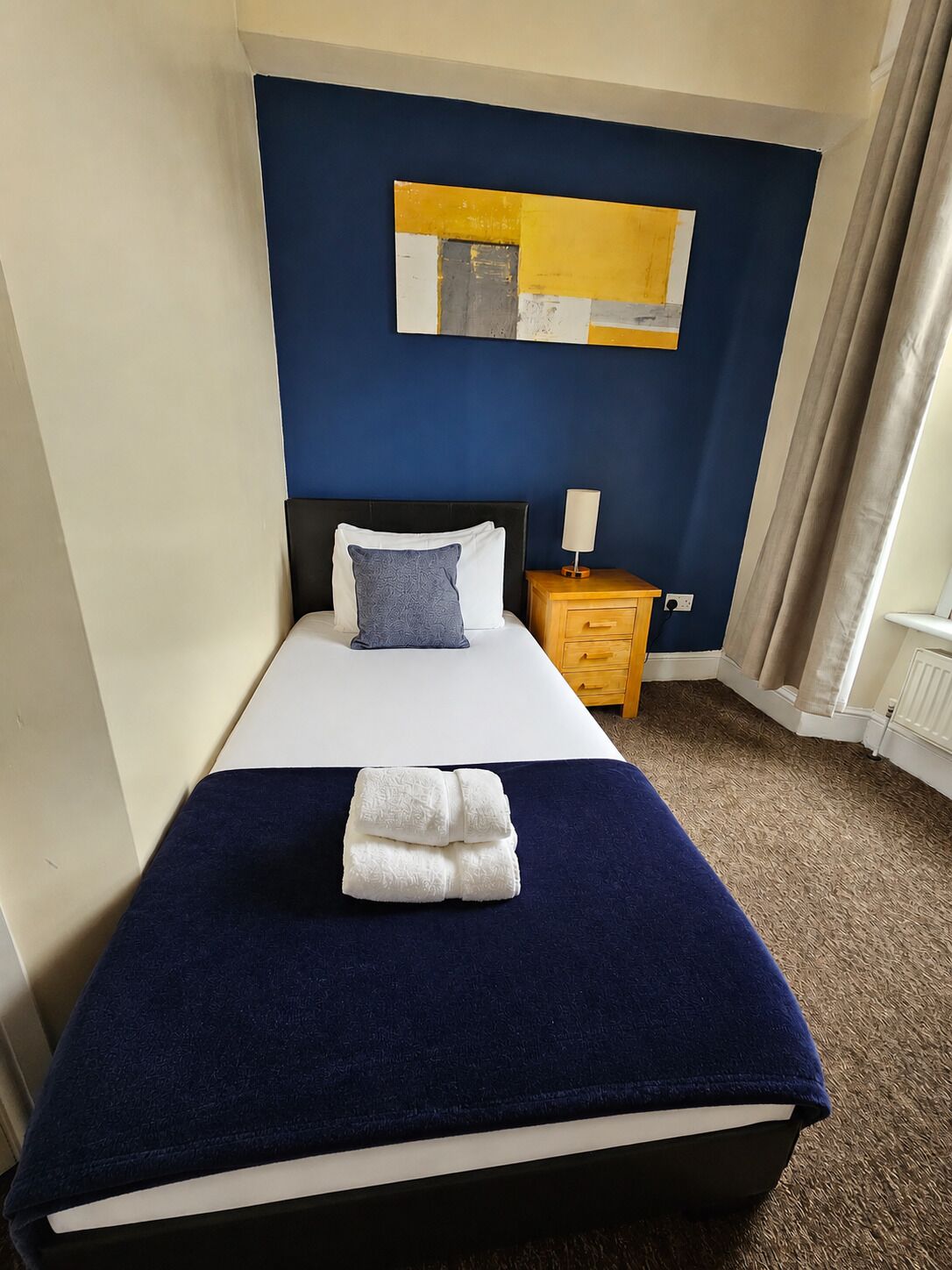A hotel room with a bed, a nightstand, a lamp, a blue wall, and a window with curtains.