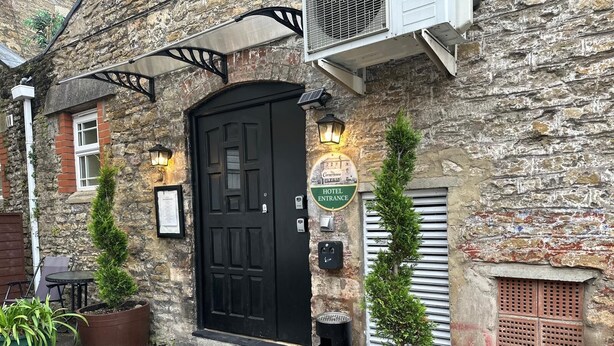 A stone building with a black door, a sign reading 'Hotel Entrance,' and two potted plants. - The Cornerhouse (Frome)