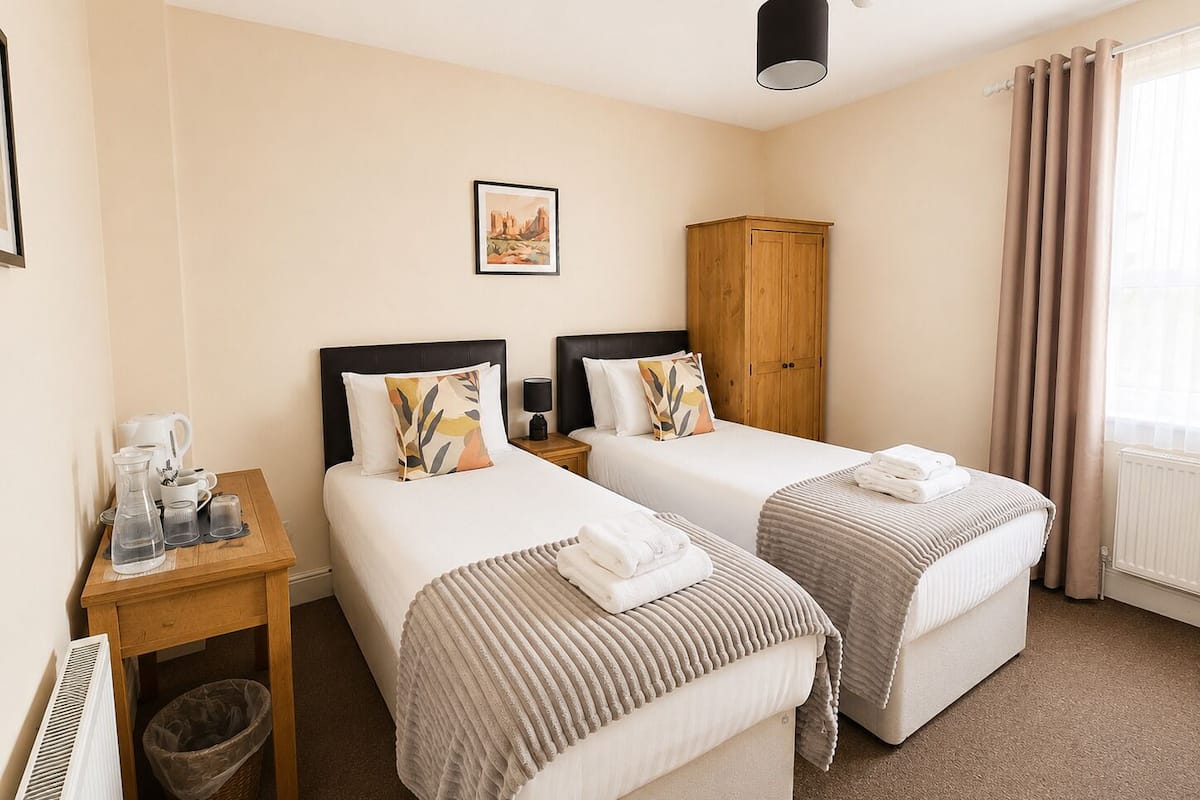 A hotel room with two beds, a wooden wardrobe, a small table with a kettle and cups, a picture on the wall, and a window with curtains.