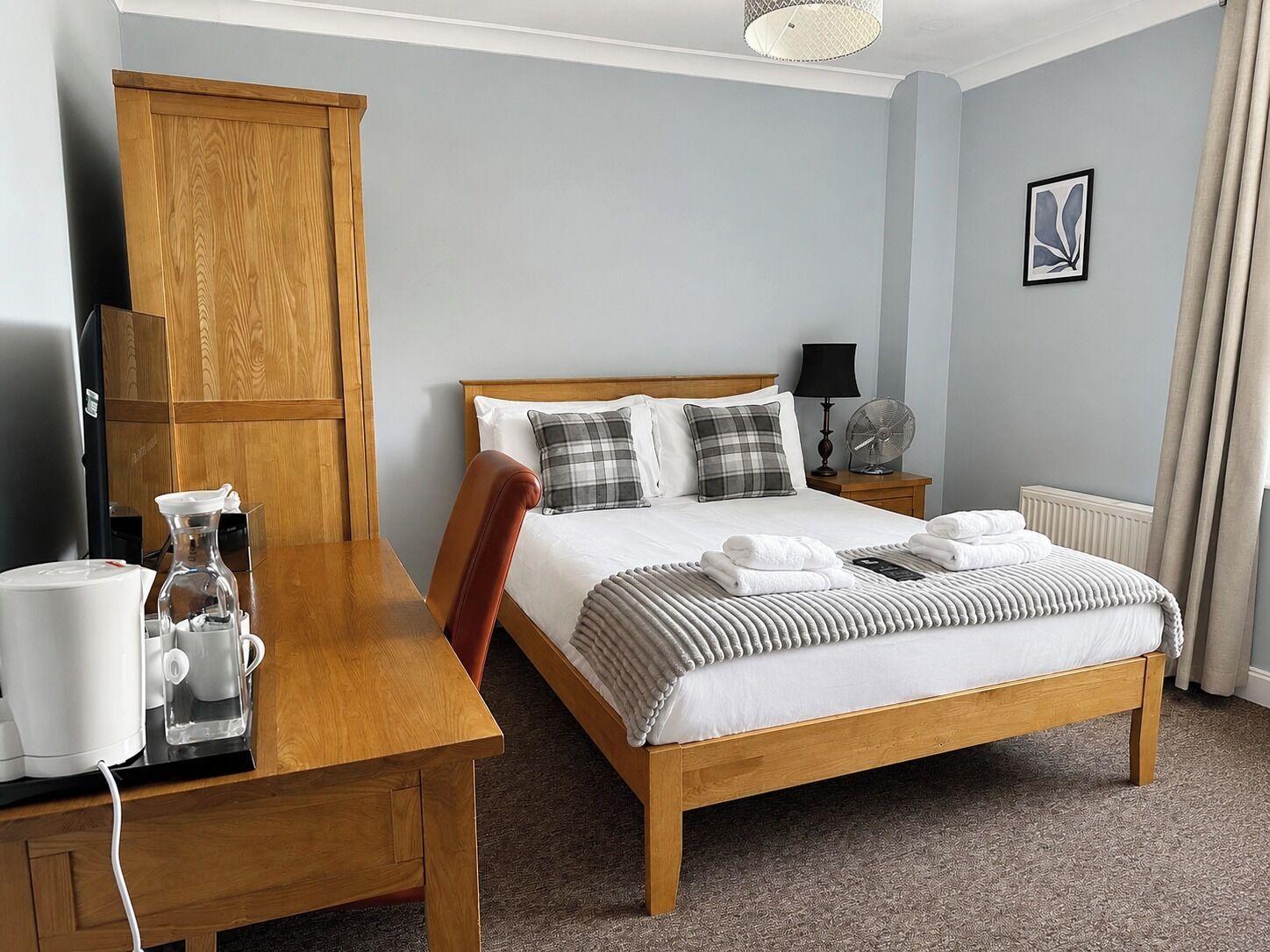 A hotel room with a wooden bed, a bedside table with a kettle, a lamp, and a fan.