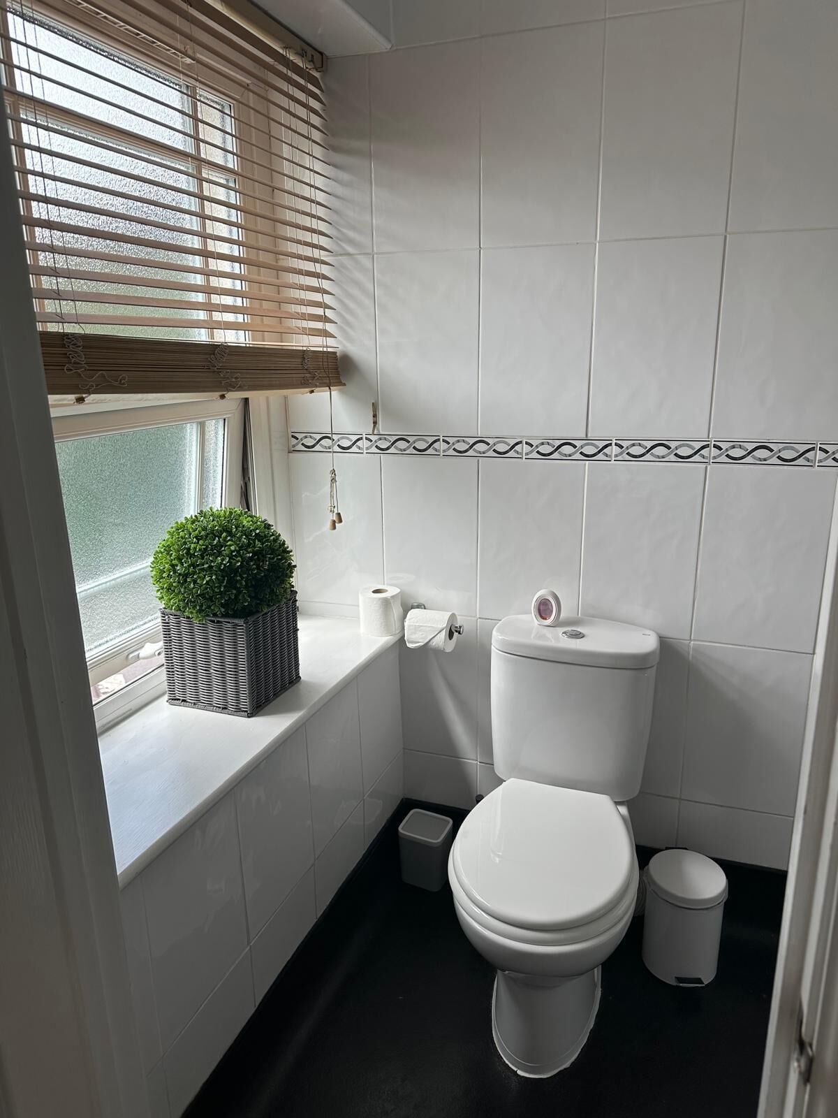 Ein Badezimmer mit Toilette, einem kleinen Mülleimer, einem Fenster mit Jalousien und einem Blumentopf auf der Fensterbank.