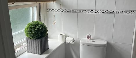 Ein Badezimmer mit Toilette, einem kleinen Mülleimer, einem Fenster mit Jalousien und einem Blumentopf auf der Fensterbank.
