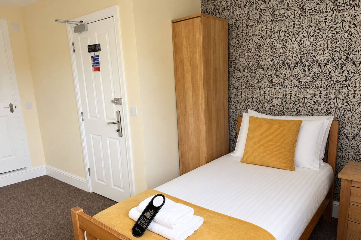 A single bed with a white and mustard-colored pillow, a wooden headboard, a wooden wardrobe, and a door with a parking sign.