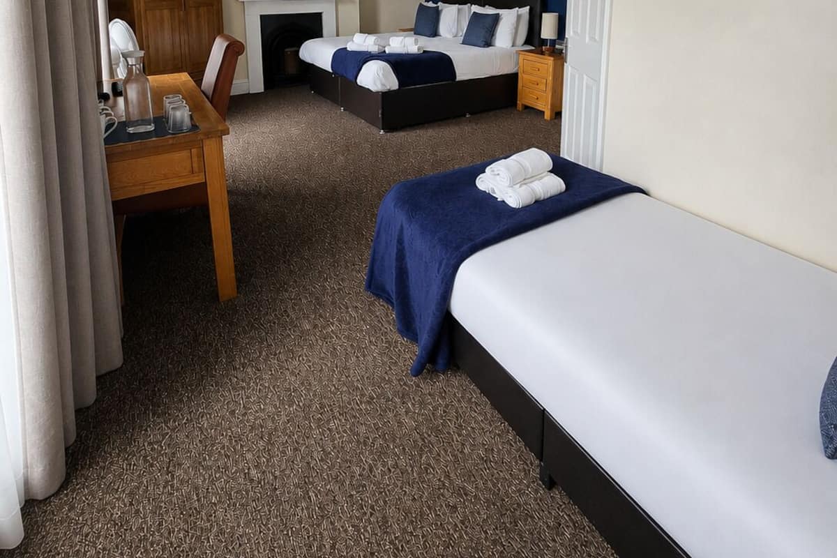 A hotel room with two beds, a desk, a chair, a fireplace, and a painting on the wall.