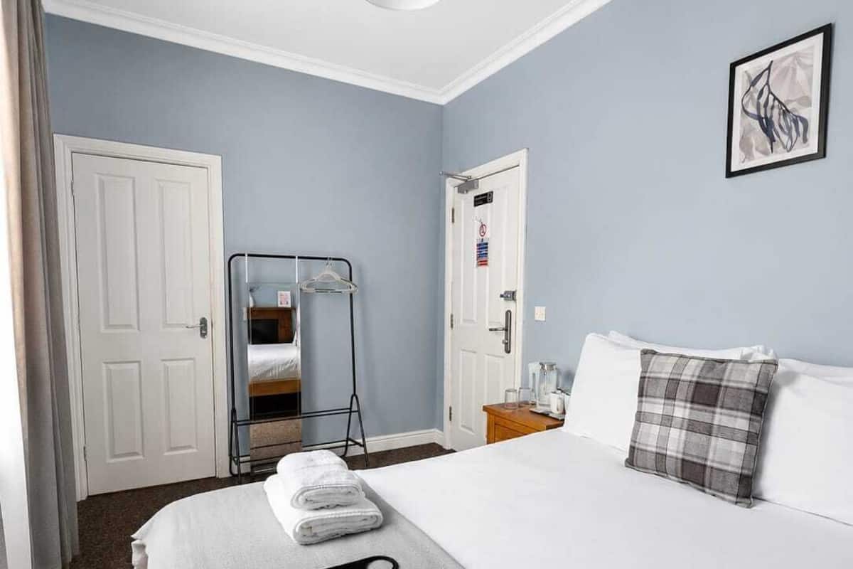 A hotel room with a bed, towels, a remote control, a painting, and a door.