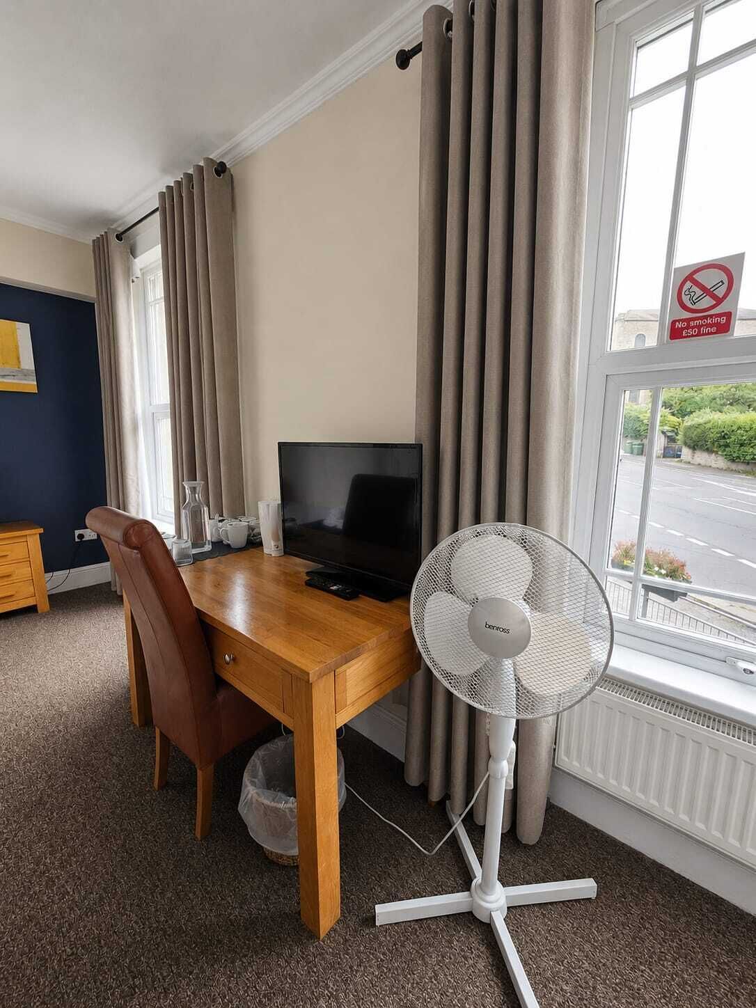 A hotel room with a wooden desk, a television, a standing fan, and a window with a view of the street.
