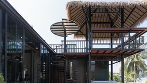 A modern building with a glass facade, a thatched-roof structure, and a staircase leading to a balcony. - Huather Villa & Cafe (Lang Suan)