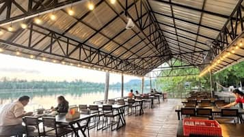 An outdoor dining area with tables and chairs, a covered roof, and a view of a lake.