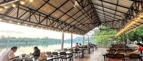 An outdoor dining area with tables and chairs, a covered roof, and a view of a lake.