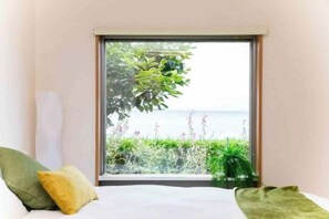 Ein Zimmer mit Bett, Kissen, einem Fenster mit Blick auf Grünflächen und ein Gewässer.