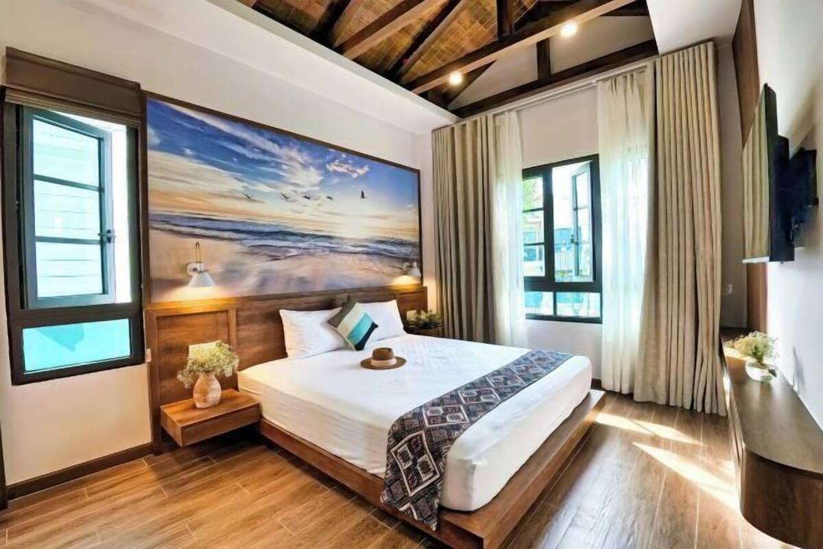 Ein Schlafzimmer mit einem großen Gemälde einer Strandansicht, einem hölzernen Kopfteil, einem Nachttisch mit einer Vase und einem großen Fenster mit Vorhängen.