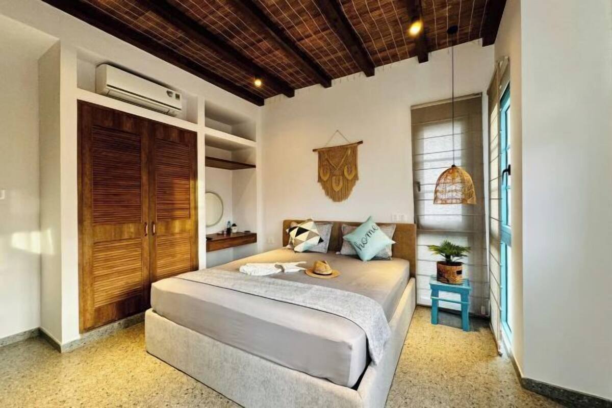 Ein Schlafzimmer mit Bett, Kleiderschrank, Nachttisch, Pendelleuchte und einem kleinen Hocker mit Pflanze.