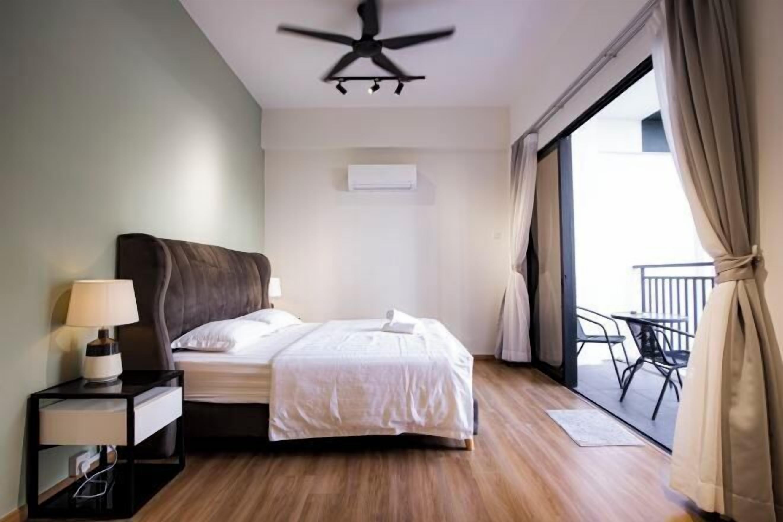 Habitación de hotel con cama, mesita de noche con lámpara, ventilador de techo, equipo de aire acondicionado y balcón con sillas.