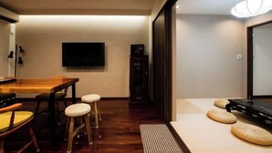 Een zithoek met een houten tafel en krukjes, een flatscreen televisie aan de muur en een traditionele Japanse tatami mat met kussens.