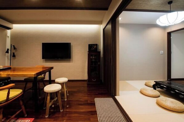 A dining area with a wooden table and stools, a flat-screen TV mounted on the wall, and a traditional Japanese tatami mat with cushions. - Kyoto Amazing The Wizarad of Oz (Kyoto)