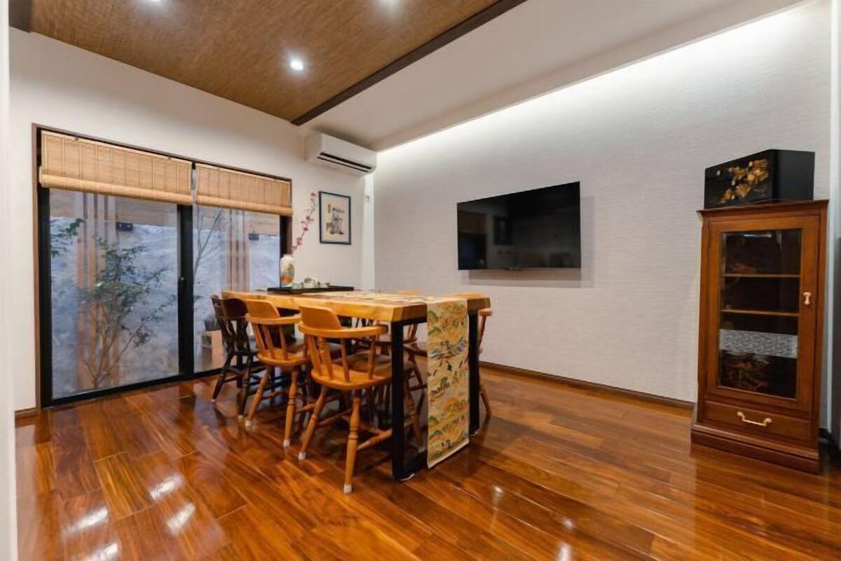 A dining area with a wooden table and chairs, a large window with bamboo shades, and a wooden cabinet.