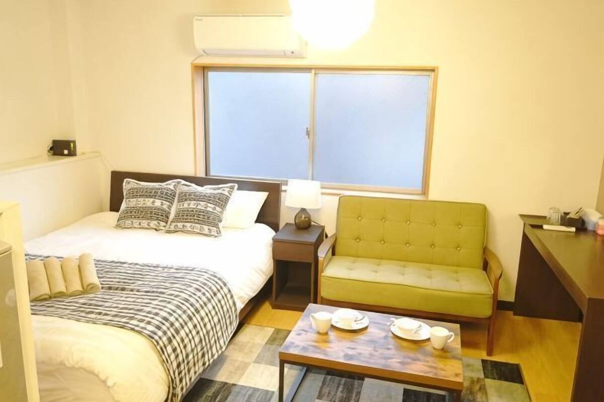 A hotel room with a bed, a green sofa, a small table with cups and saucers, a nightstand, and a window.