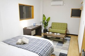 Une chambre d’hôtel avec un lit, un bureau, un fauteuil vert, une petite table et une plante en pot.