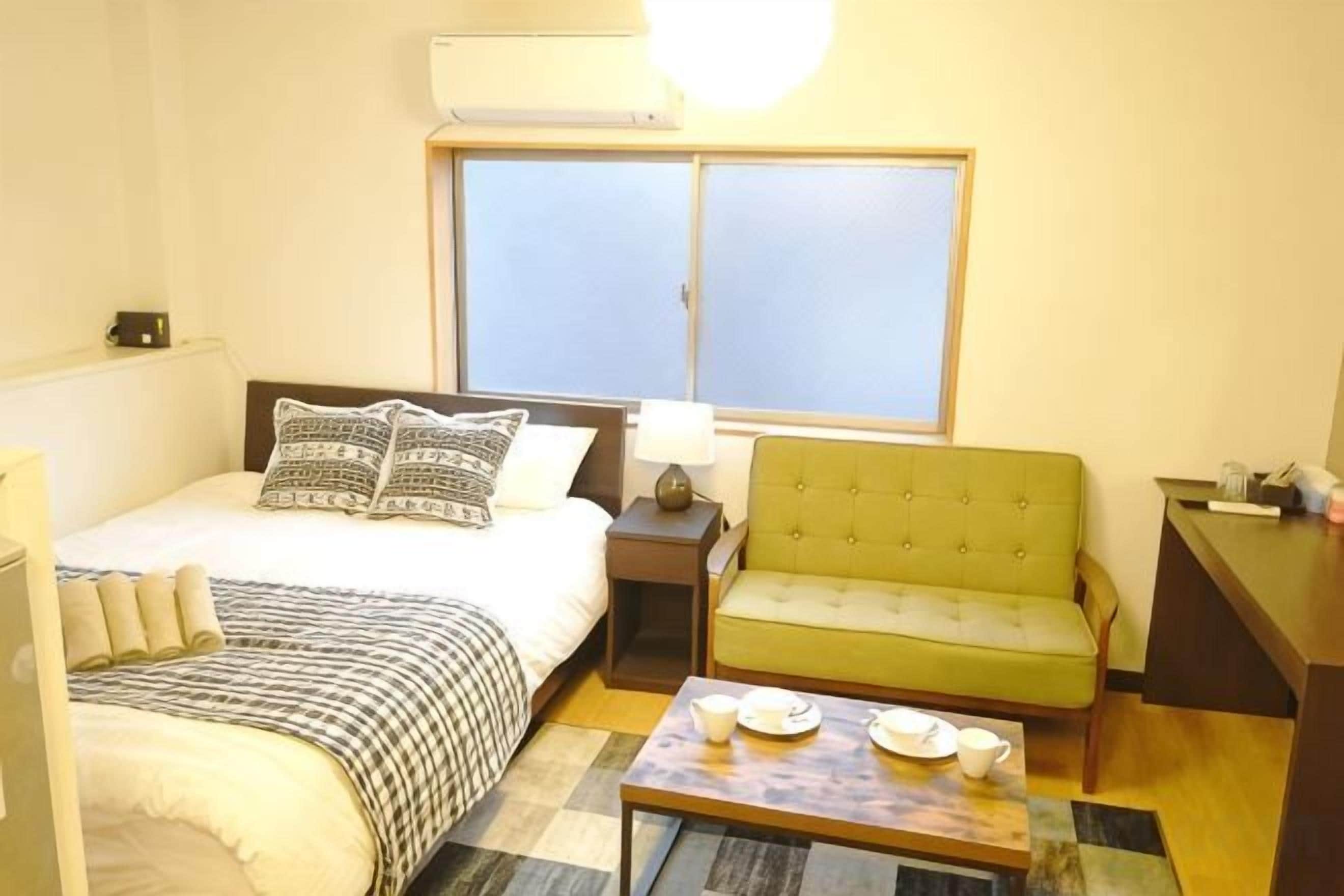 A hotel room with a bed, a green sofa, a small table with cups and saucers, a nightstand with a lamp, and a window with blinds.