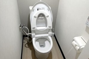 A white toilet with a bidet attachment, a toilet paper holder, and a wall-mounted flush button. - YANG house (Osaka)