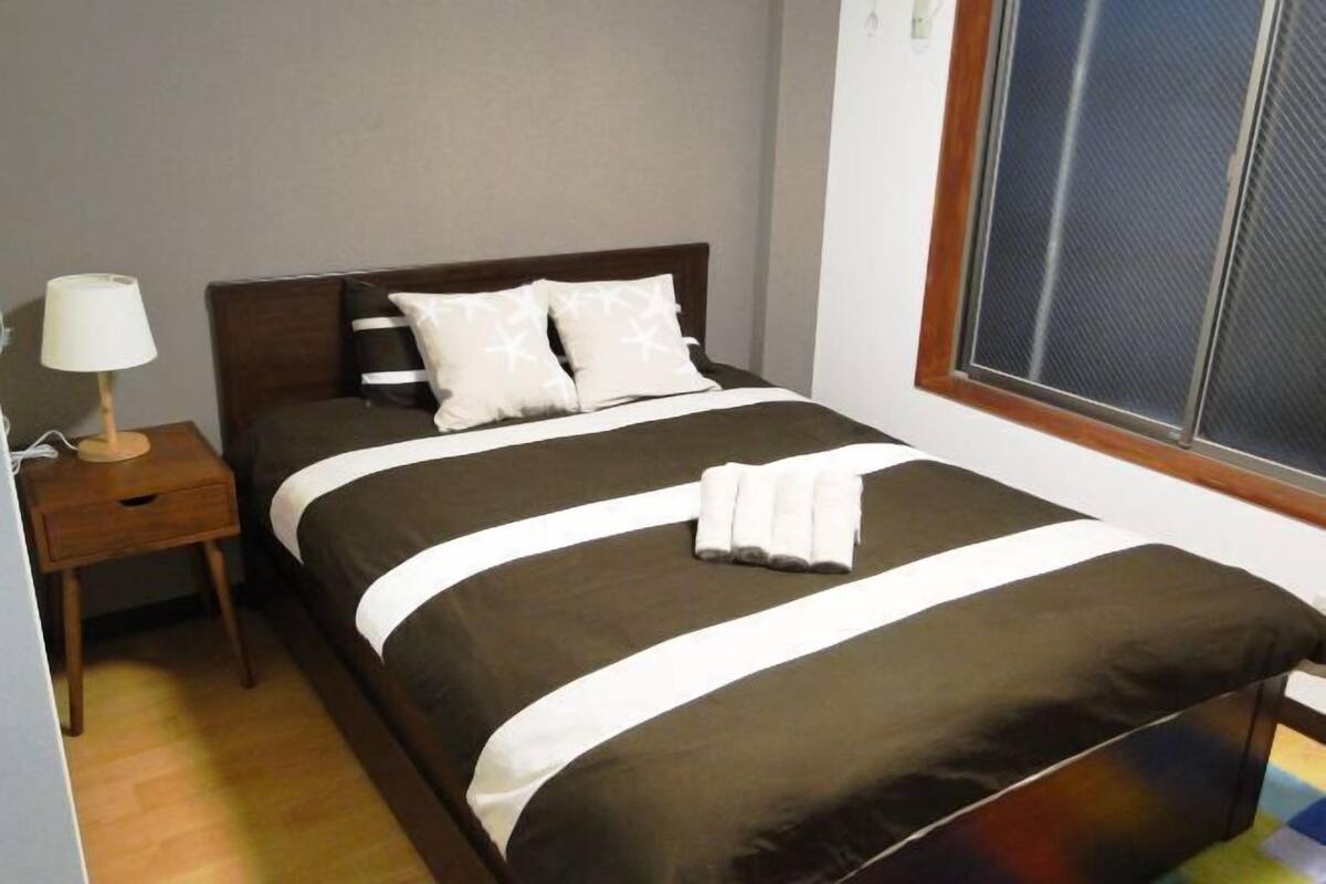 A neatly made bed with a dark and white striped bedspread, a wooden nightstand with a lamp, and a window with a view.