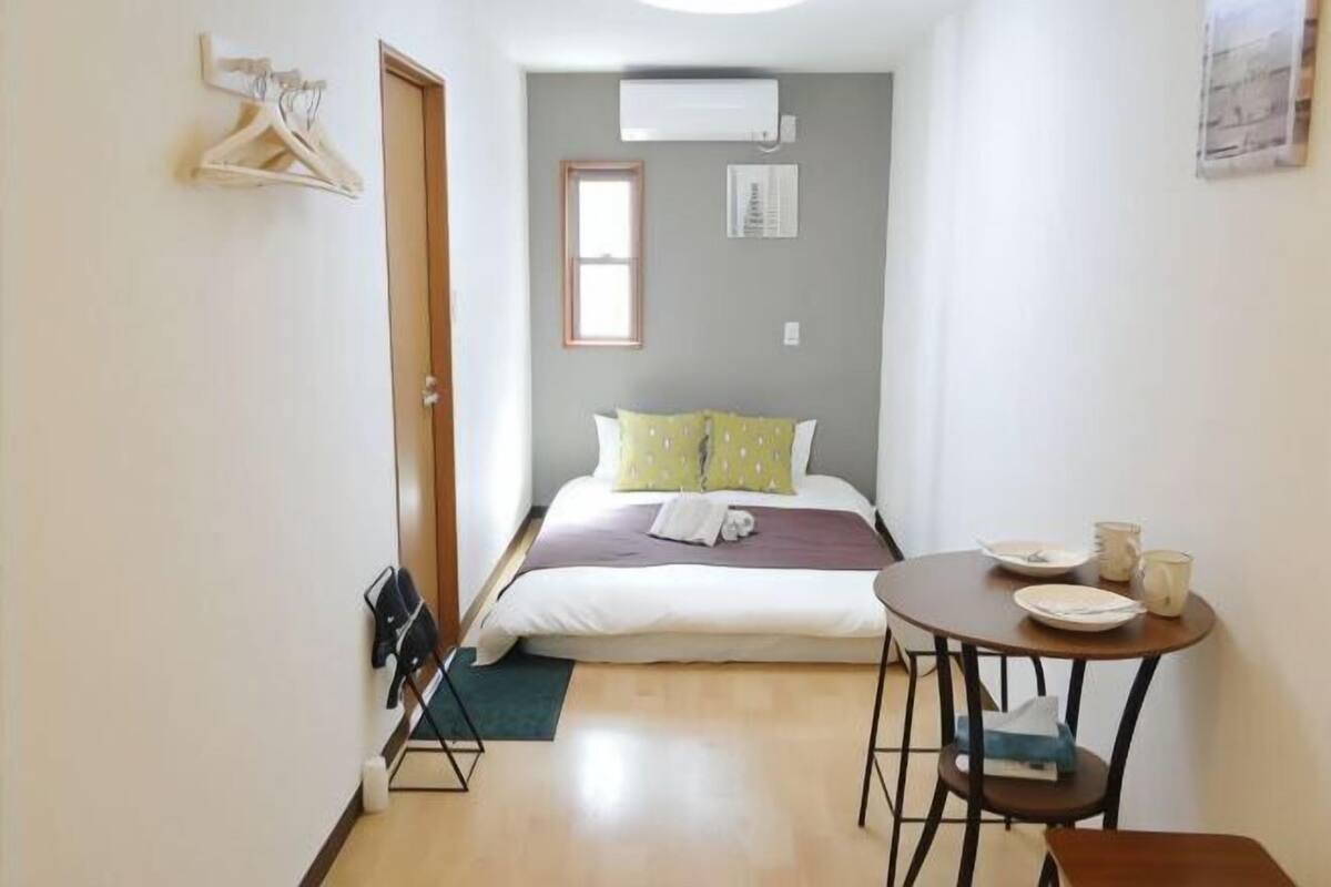 A small, neatly arranged bedroom with a bed, a table with plates and cups, a hanging coat rack, and an air conditioning unit on the wall.