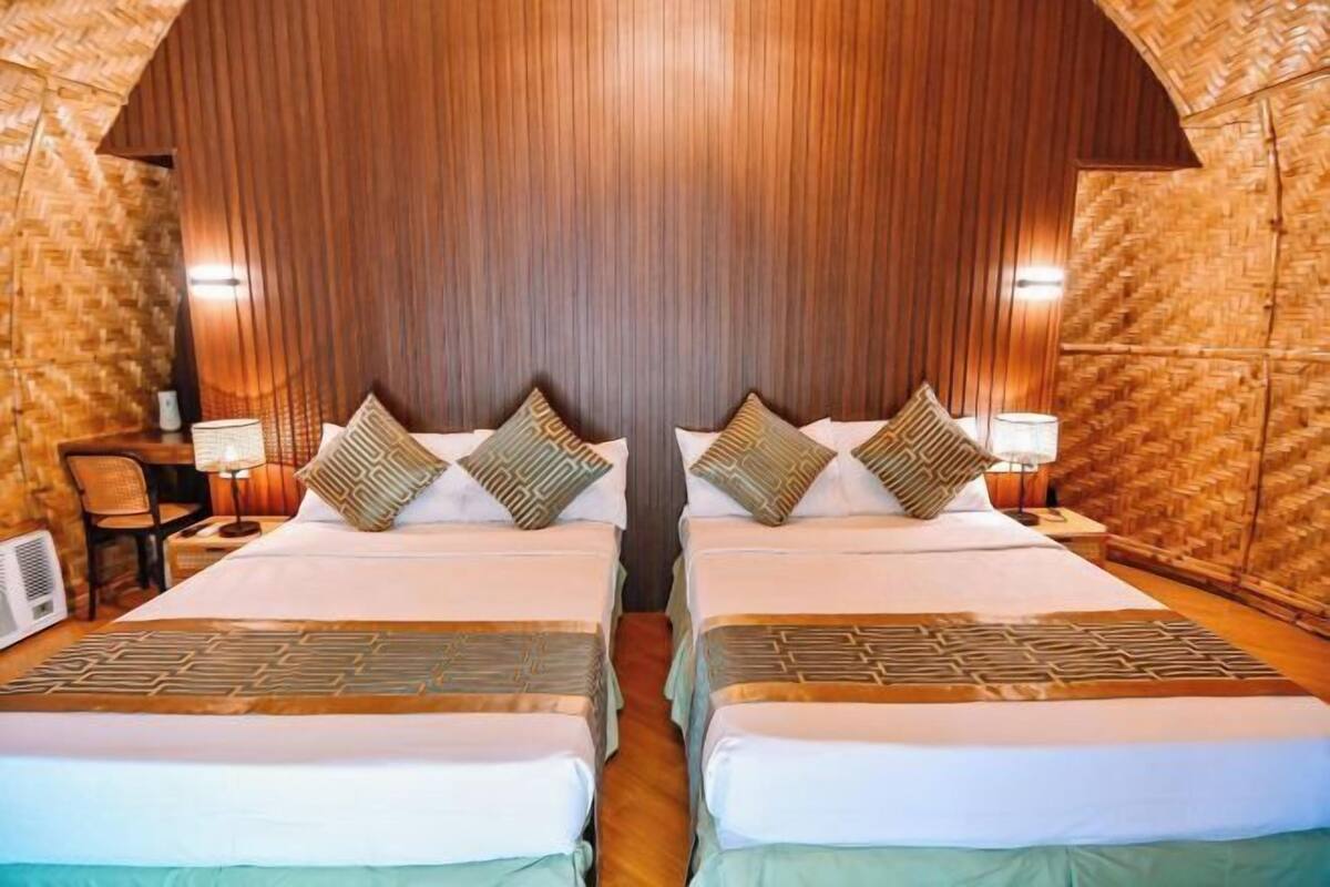 A double room with two beds, each with patterned bedding and pillows, a wooden headboard, and a small table with a lamp.