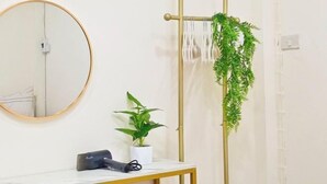 Une salle de bain avec un miroir rond, une petite table avec un sèche-cheveux, une plante en pot et un support vertical pour plantes avec des feuilles vertes.