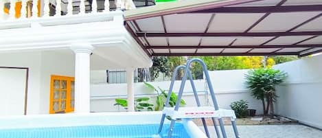 A swimming pool with a ladder, a covered patio, and a white building with a balcony.
