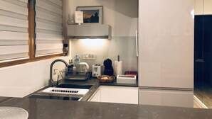 A modern kitchen with a sink, countertop, and a built-in oven. There are shelves above the counter and a window with blinds. - INNA LUXE (Belgrade)